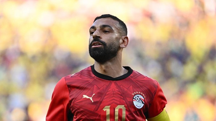 Bearded male soccer player in a red jersey with number 10 on the field, yellow armband visible on the arm, out-of-focus crowd behind.