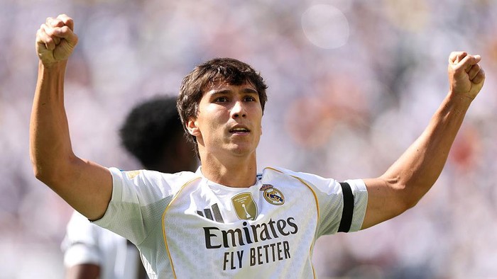 Soccer player in white Real Madrid kit raises arms in triumph on the field at a match.