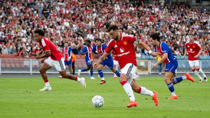 Soccer match with players in red and blue uniforms chasing the ball on a green field, crowded stands in the background.