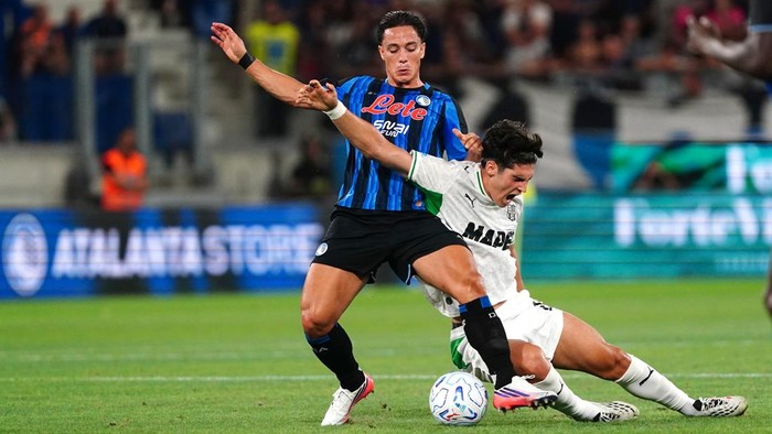 Soccer players contest the ball during a match, one in blue-and-black stripes and the other in white.