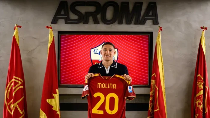 Soccer player stands in front of a crest on a screen, holding a red jersey with the number 20 and the name Molina, flanked by red flags under a sign that reads ASROMA.