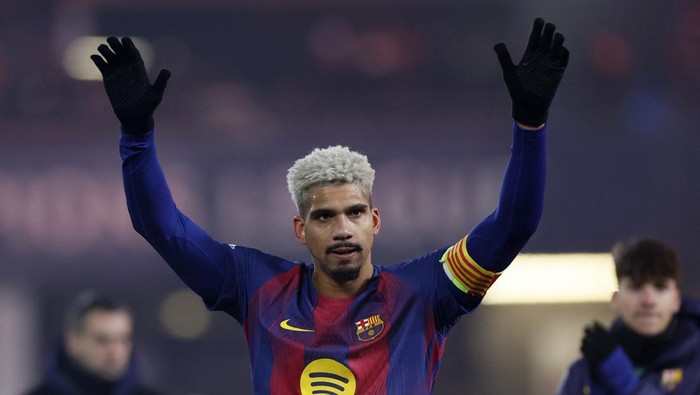Footballer with bleached hair raising both arms in celebration on the pitch, wearing a Barcelona jersey and captain's armband in yellow-red stripes