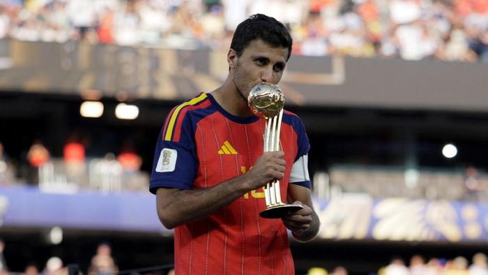 Soccer player in a red-and-blue jersey kissing a tall gold trophy on the field.