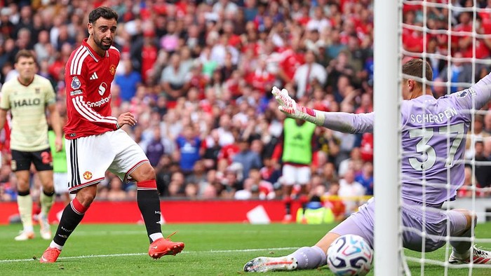 Manchester United player in red shoots toward goal as goalkeeper dives to block the shot from close range amid a crowded stadium crowd.