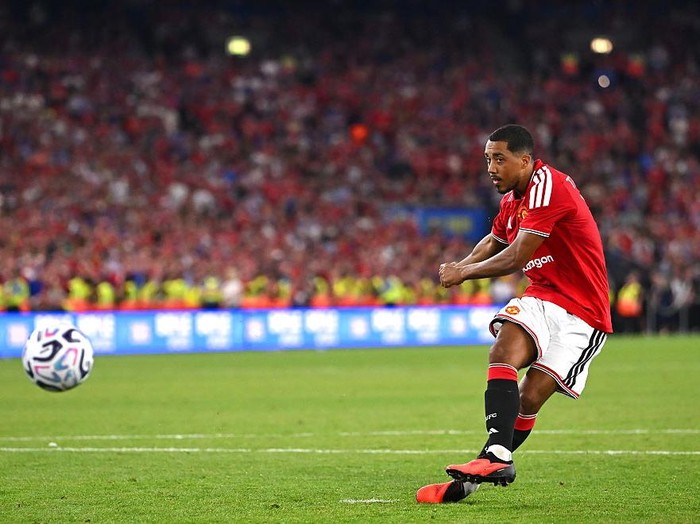 Soccer player in a red jersey kicking a ball on a green field during a match with a packed stadium in the background, fans visible.