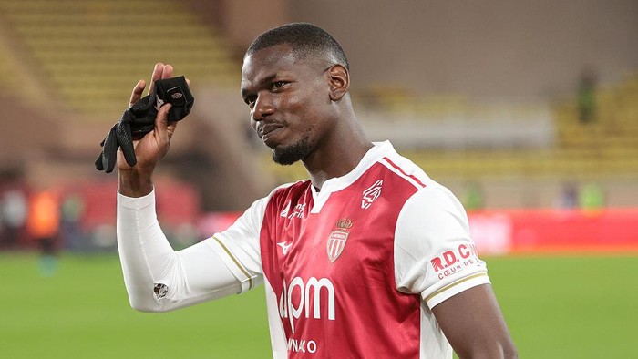 Soccer player in a red-and-white jersey lifts his gloved right hand on a football pitch with blurred stands in the background.