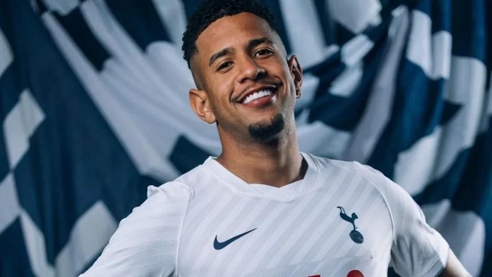 Smiling man in a white Tottenham Hotspur jersey against a blue abstract background.