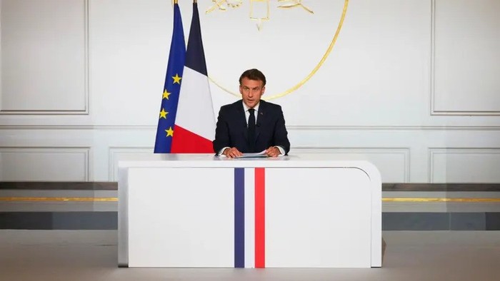 Male official in a dark suit sits at a white podium with French flag colors, flanked by the French and EU flags in a formal gov’t chamber.