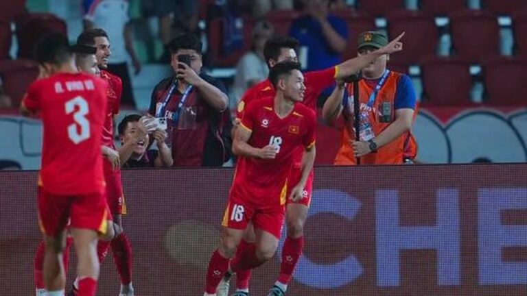 Soccer players in red uniforms celebrate near the sideline; one runs forward with a smile while another points, with fans and a photographer in the background.