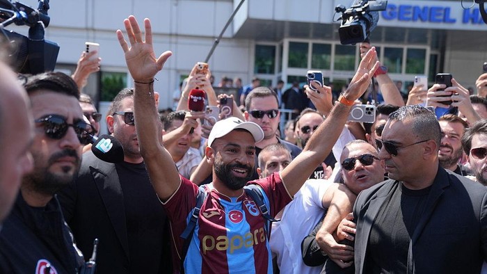 Athlete in a maroon and blue jersey raises both arms in celebration as fans and cameras surround him outside a building.