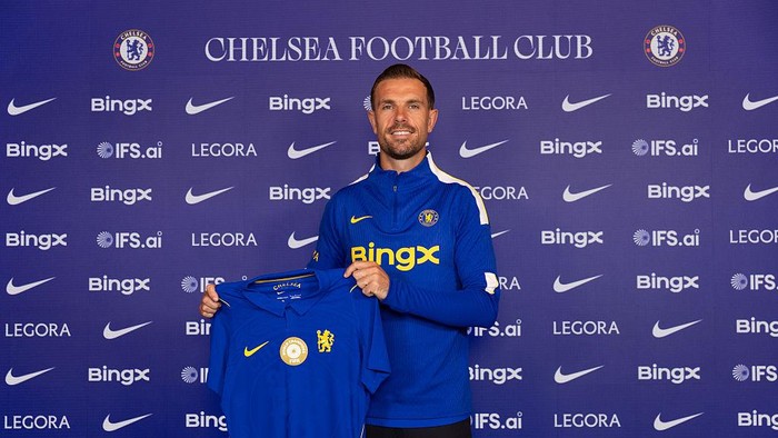 Male Chelsea FC staff member in a blue training top holds up a blue club jersey at a signing event, backdrop shows Chelsea and sponsor logos.