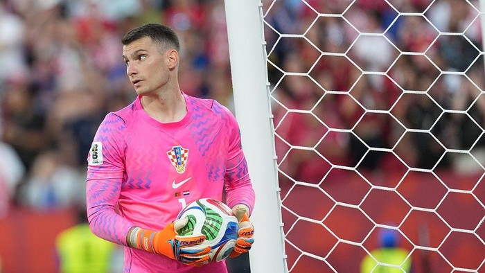 Goalkeeper in a pink kit and orange gloves holding a soccer ball beside the goal net, looking to the left.