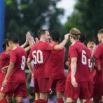 Soccer players in red jerseys celebrate with a group high-five on the field, a referee in yellow watches nearby.