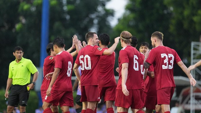 Soccer players in red jerseys celebrate with a group high-five on the field, a referee in yellow watches nearby.