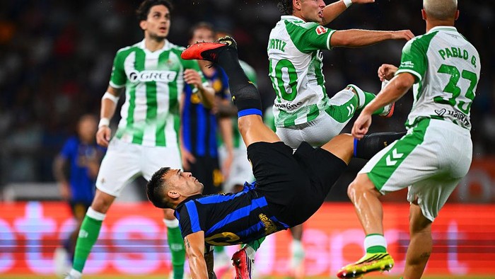 Soccer match action: a blue-and-black kit player performs a mid-air bicycle kick while green-and-white team players surround him.