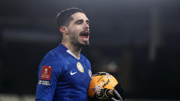 Chelsea goalkeeper in a blue kit holds a yellow ball and shouts on the field, FA Cup patch visible on the sleeve with 'No room for racism' badge nearby.