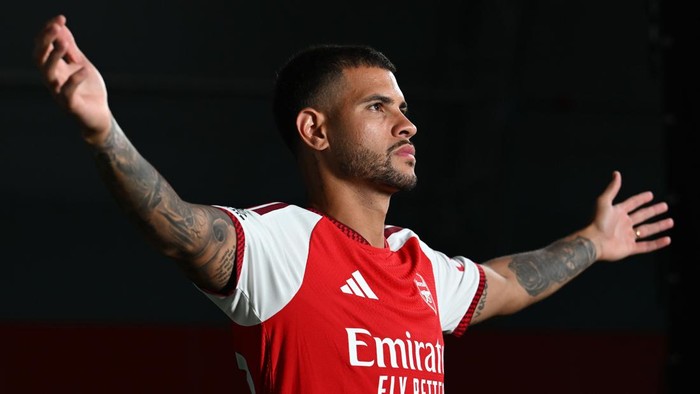 Footballer in Arsenal jersey with arms wide, celebrating on the pitch in a red kit with tattoos visible on his arms.