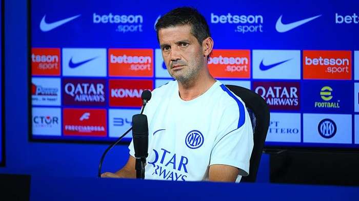 Man in a white Inter Milan training shirt sits at a press conference microphone, with a blue sponsor-filled backdrop behind.