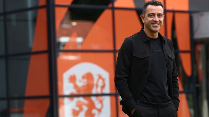 Smiling man in a black jacket stands with hands in pockets outside a glass-fronted building with orange panels and a white logo behind him.
