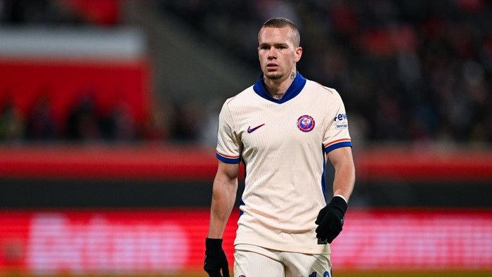 Male footballer in a light beige kit with blue trim on a stadium field, wearing black gloves and looking ahead.