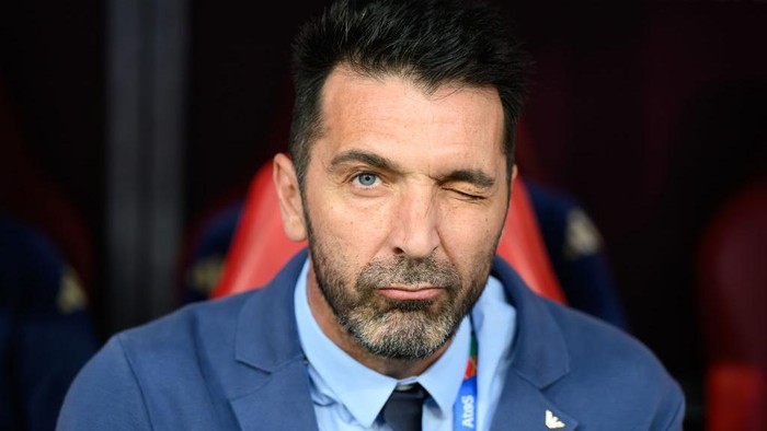 Man in a blue blazer winking at the camera while seated on a bench in a stadium setting.