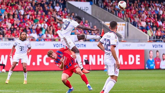 Action shot of a soccer match: a player in white jumps to head the ball while an opponent in red and blue is toppled on the ground, with other players watching on green field and advertising boards in the background.