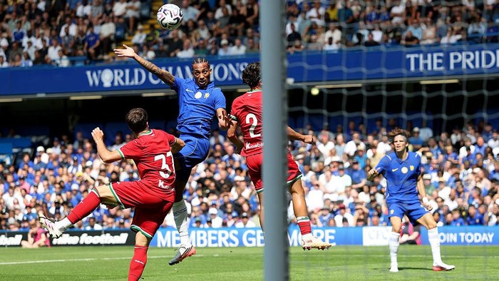 Soccer players in blue and red contest a high ball near the goal as a blue striker jumps to head it, with a crowded stadium in the background.