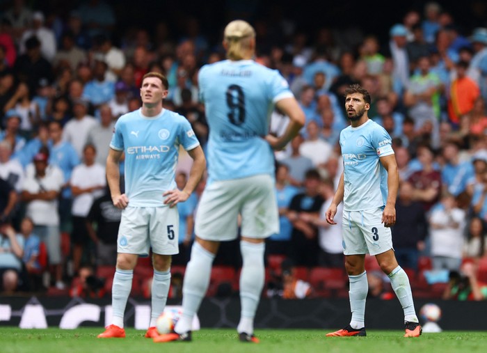 Manchester City players in light blue kits stand on the pitch during a game, with a crowded stadium in the background.