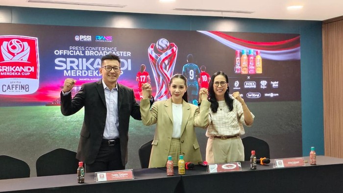 Three individuals at a press conference table raise their fists in solidarity, with a banner and sponsor logos visible in the background.