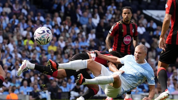Soccer players battle for the ball during a match; a light-blue jersey player slides in while opponents in black-and-red contest nearby.