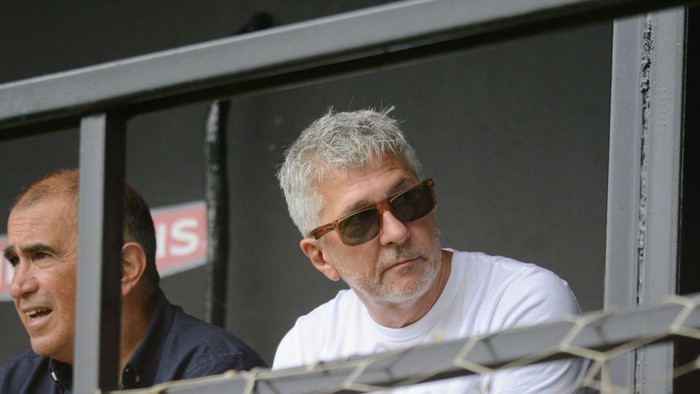 Two men seated in a stadium railing, the foreground man with gray hair wearing sunglasses and a white shirt, watching intently.