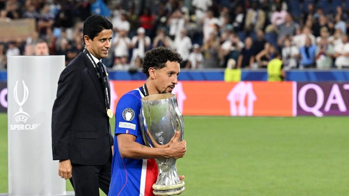 Soccer player in blue holds a large trophy on a stadium field, with a man in a suit beside him and cheering crowd in the background.