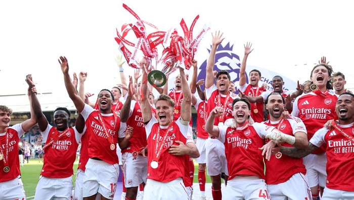 Arsenal players celebrate on the field with a trophy and red ribbons, cheering and raising their hands in victory.