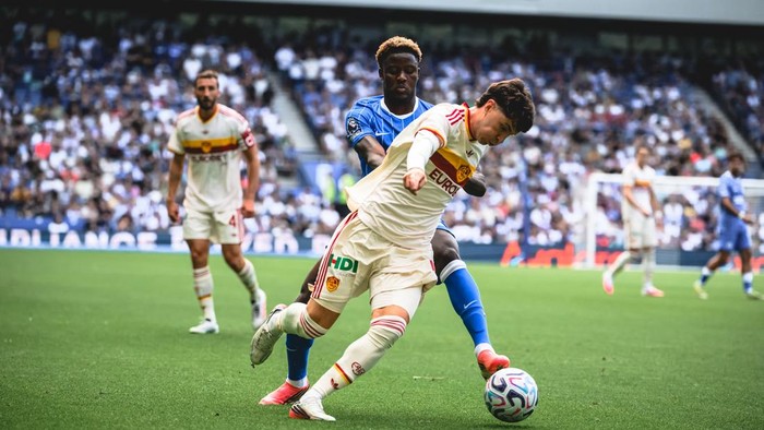 Soccer players in cream and blue compete for the ball on a green field with a crowded stadium in the background, one player kicking the ball while others watch.