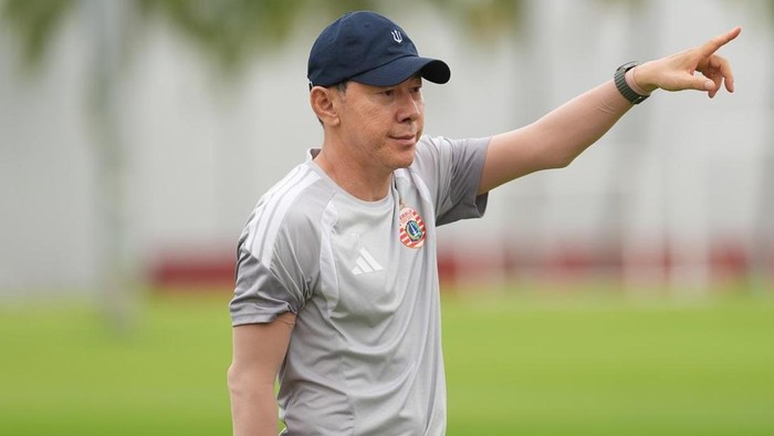Coach in a gray athletic shirt and navy cap points to the right on a grassy field during training.