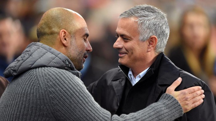Two men in jackets warmly greet each other with a handshake and smile on a stadium sideline.