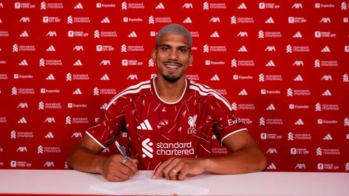 Liverpool FC player signs a contract at a press event, wearing a red kit with sponsor logos behind him.