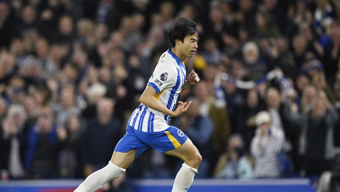 Soccer player in blue-and-white striped kit runs with the ball during a match, stadium crowd blurred in background.