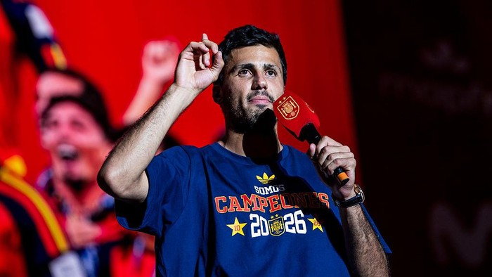 Man speaks into a microphone on stage, wearing a blue 'Campeones 2026' shirt, with a red background and cheering crowd behind him.