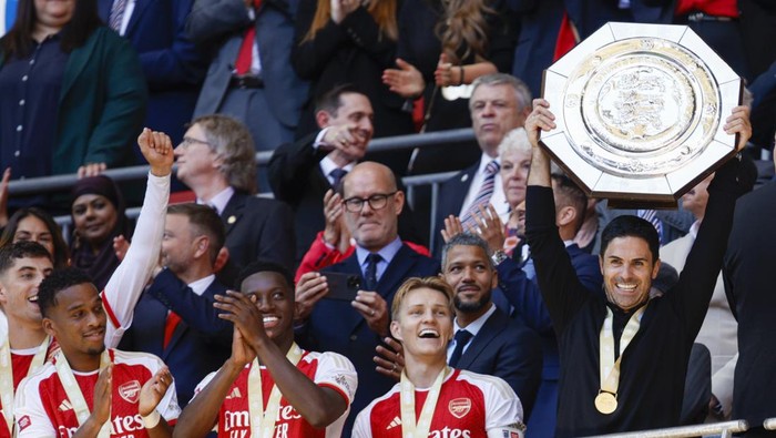 Arsenal players celebrate winning a championship, with the goalkeeper lifting a large ornate shield trophy as teammates cheer nearby.