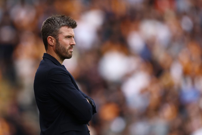 A man in a dark blazer with arms crossed, watching a game on the sideline as blurred stadium crowds glow in the background.
