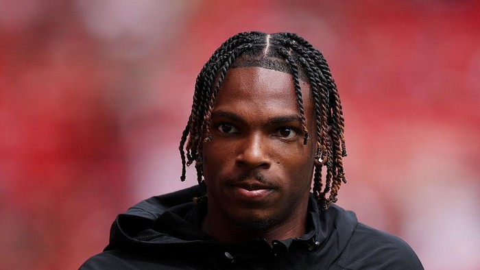 Close-up portrait of a man with braided hair wearing a black jacket, looking straight ahead with a neutral expression against a blurred red background.