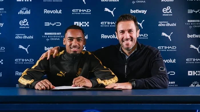 Two men sit at a signing table, smiling, with a backdrop of sponsor logos behind them (sports deal signing)