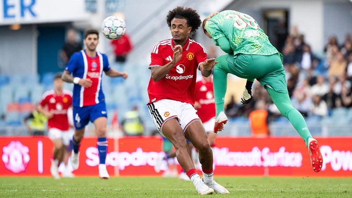Soccer player in a red jersey challenges a green-clad goalkeeper as the ball flies nearby on a sunny pitch during a league match.