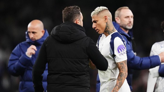 Two men greet each other with a handshake on a soccer field; one in a white kit with visible tattoos on his arm, the other in a black jacket.