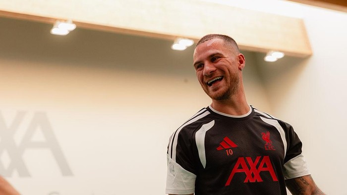 Smiling footballer in a black Liverpool training shirt with AXA sponsor indoors.