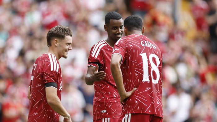 Three soccer players in red uniforms celebrate on the field; one faces the camera smiling while the other two show their backs, including number 18 with the name GAKPO.