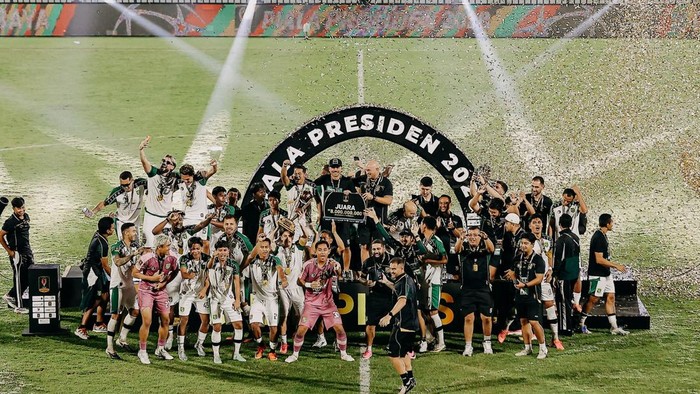 A soccer team celebrates their victory on the pitch, lifting trophies under a circular winner’s arch with confetti spraying down.