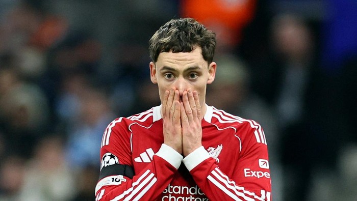 Soccer player in a red Liverpool kit covers mouth with hands in surprise on the field.