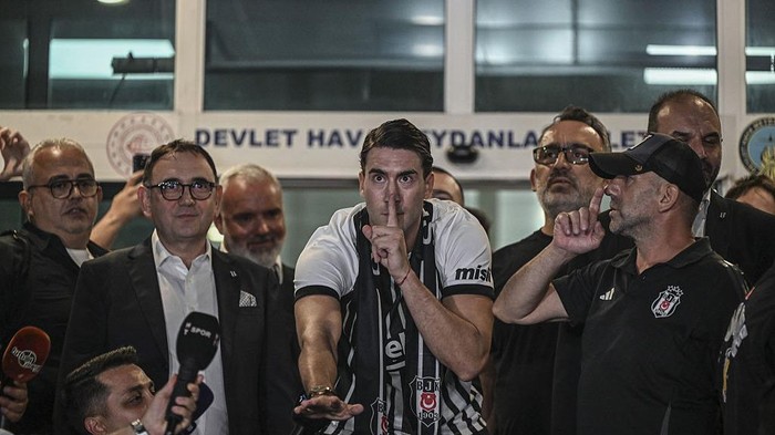 Football player with a black-and-white scarf holds a shushing gesture while surrounded by reporters and officials at a press event.
